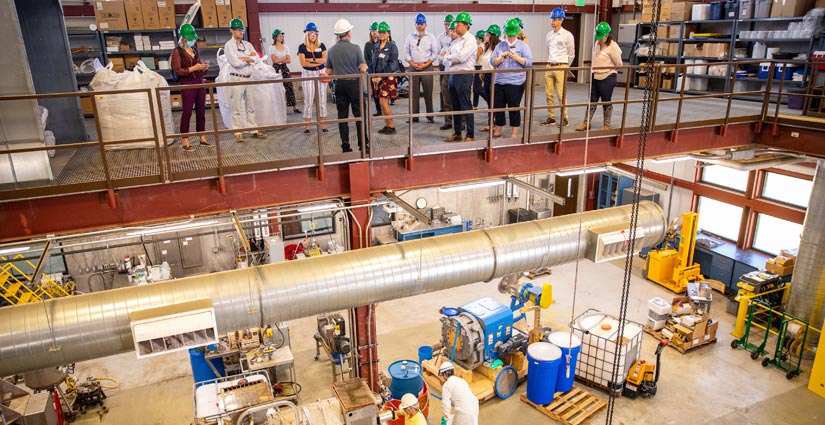 View of a two-story facility, with workers and equipment on the bottom floor and visitors in hard hats on the top floor.