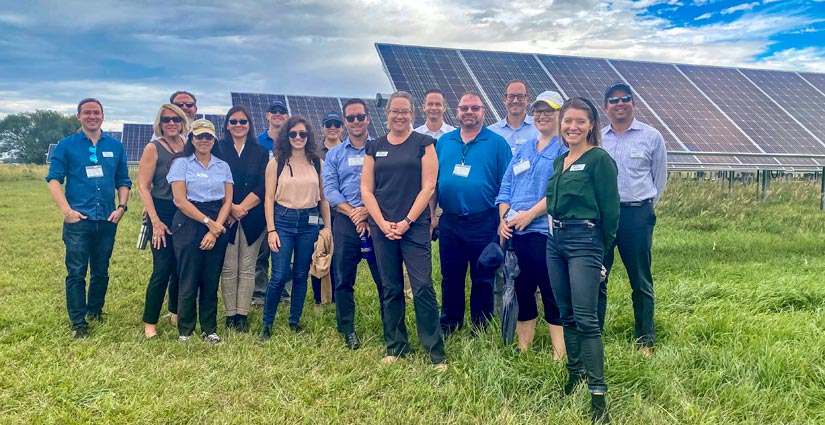 Sixteen people smile and pose on a grassy field in front a set of solar panels.