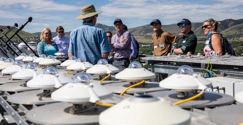 A person with their back to the camera stands among electrical equipment outdoors while speaking to a group.