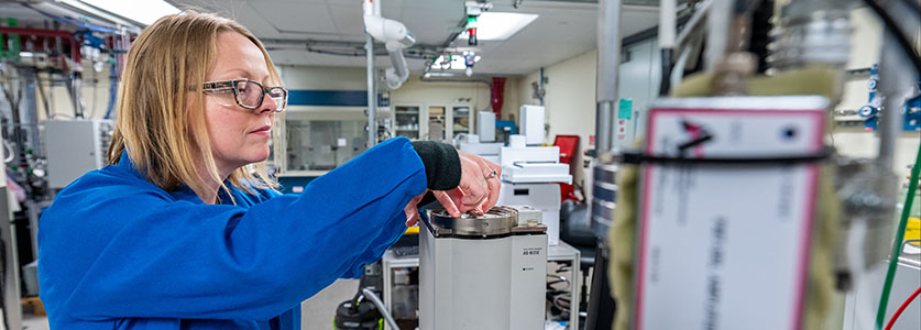 One researcher with safety glasses in a lab coat working on equipment in a lab. 