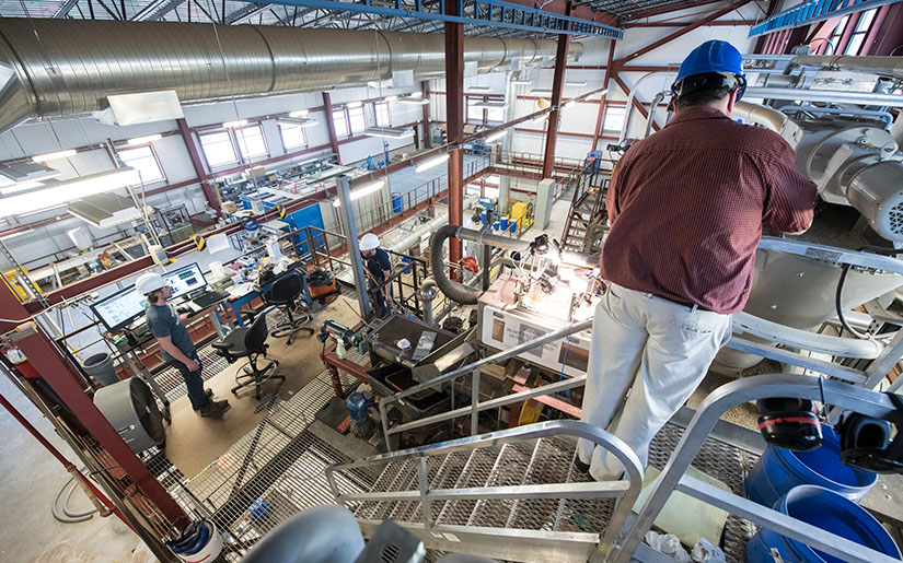 One researcher in a hard hat working on equipment in a lab.