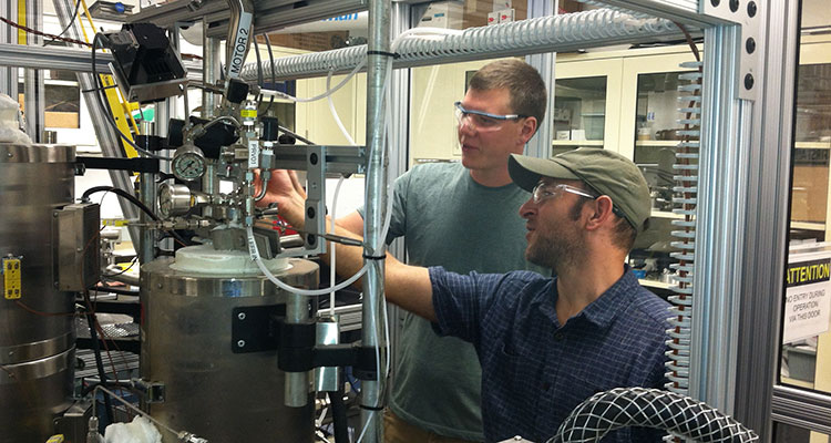 Photo of two engineers in safety glasses working on a reactor system comprised of metal tanks, tubing, pipes, and wires, which is used to investigate in situ and ex situ catalytic fast pyrolysis and can be used to generate bio-oil for compositional analysis.