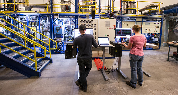 Photo of two engineers with their backs to the camera, standing in front of tall tables with laptops and computers that are sitting in front of the large Davison circulating riser, which is a two-level set of multi-colored, metal scaffolding, pipes, tubes, and hoses that connect to several control panels with dials and monitors.