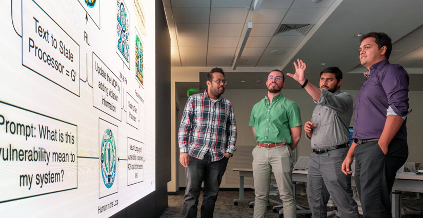 Four people stand in front of a wall bearing a presentation slide; one speaks and gestures toward the slide.