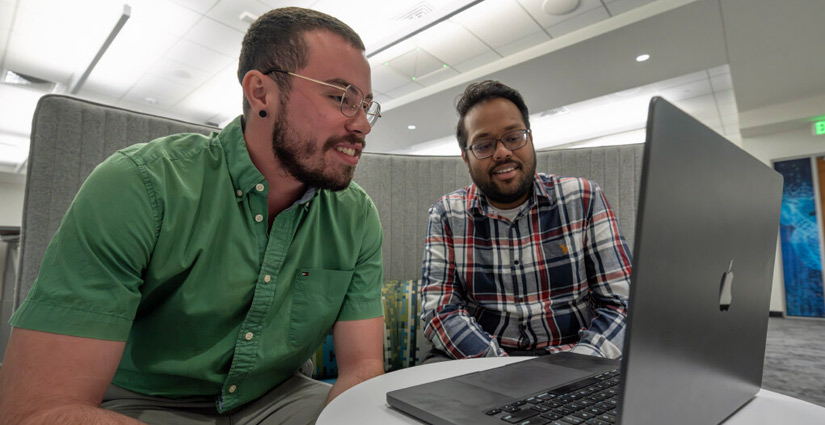 Two people sit in a booth together, looking at a laptop screen.