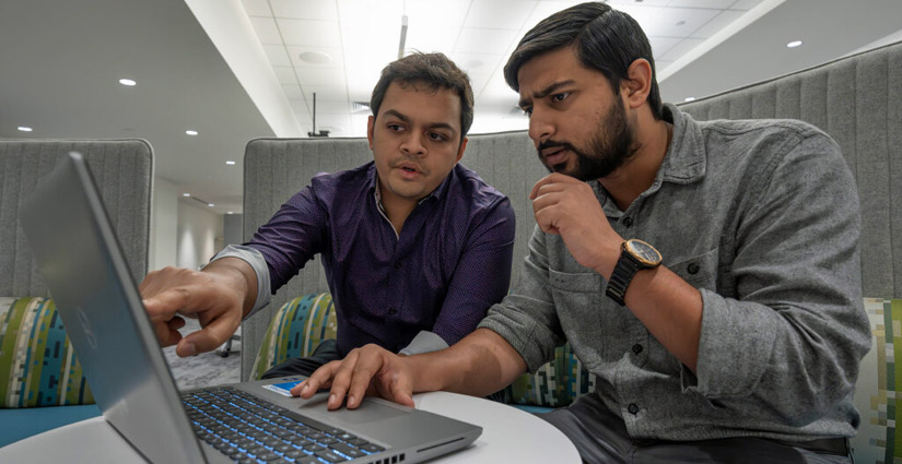 A person seated alongside another in a booth points at the laptop screen both are viewing.