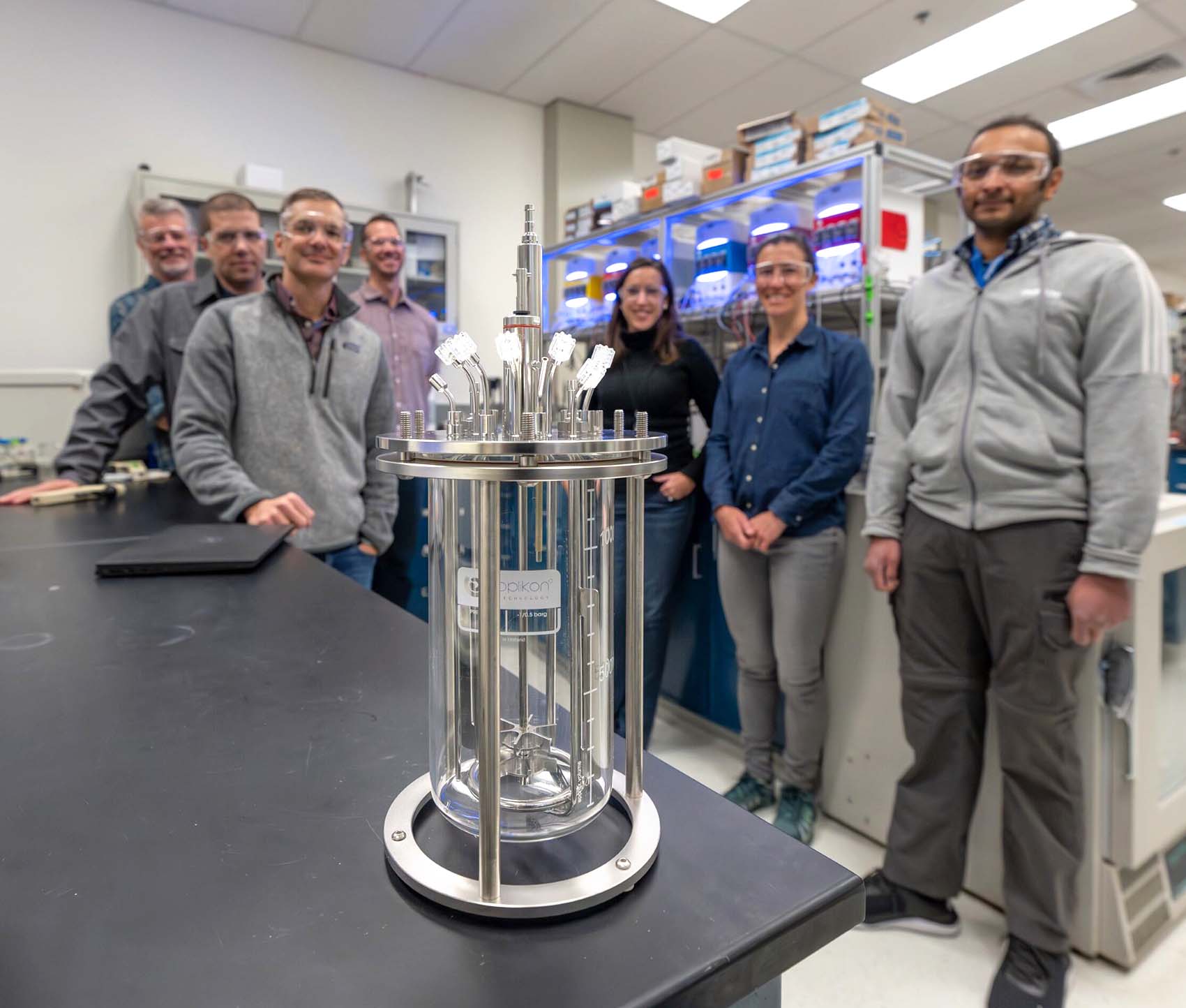 A group of seven individuals stands in a laboratory behind a cylindrical device.