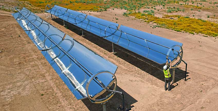 A man in a hardhat and reflective vest works on one of two reflective parabolic troughs amidst a desert landscape.
