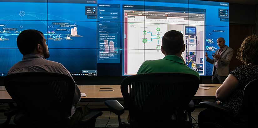 Photo of three people at a desk listening to a presentation from a man standing in front of a wall of computer screens. On the screens are visualizations of NREL's cybersecurity capabilities and data.