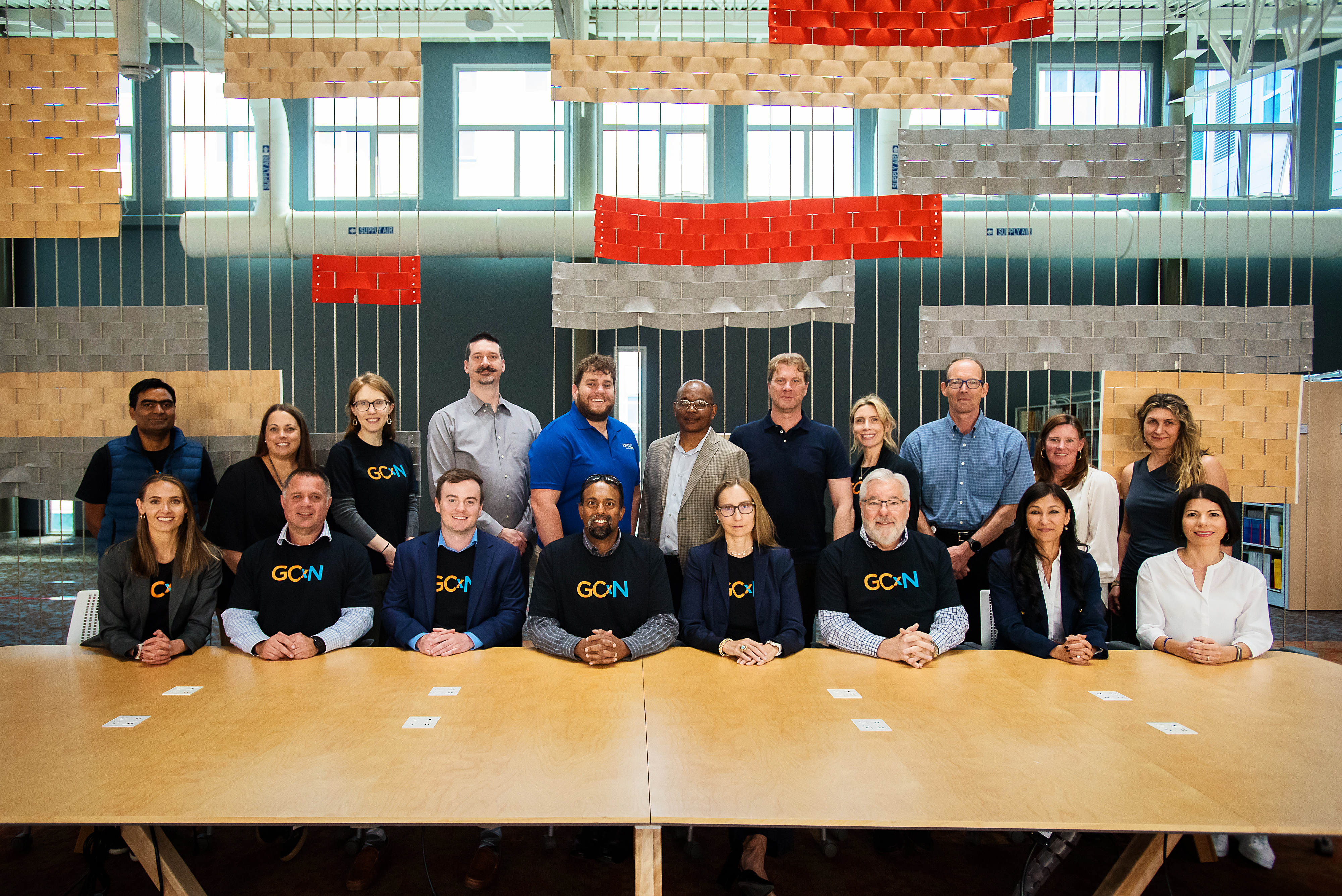 A group of people sitting at a long table wearing t-shirts that say GCxN