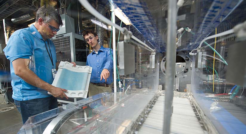 Both wearing eyewear and blue shirts, two researchers communicate while standing on the side of a long piece of machinery, one holding a blue and white object while the other points to the machinery.