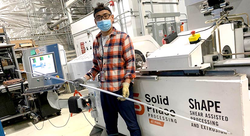 A person wearing a mask holds two long tubes standing in front of ShAPE-labeled processing machinery