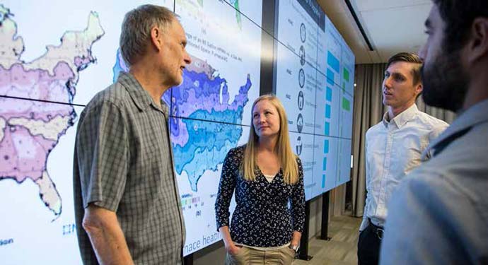 A group of people stand in front of screens with maps and graphics displayed across them.