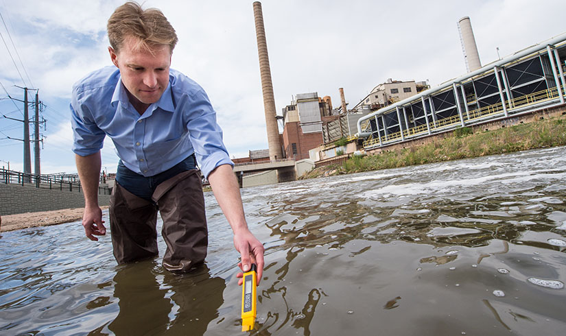 A man in a dress shirt standing in front of a power plant gets a temperature reading and water samples from a body of water.