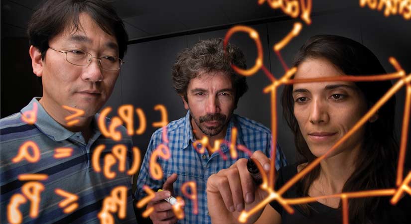 A photo of a group of people writing equations in orange marker on a clear, plastic board. 