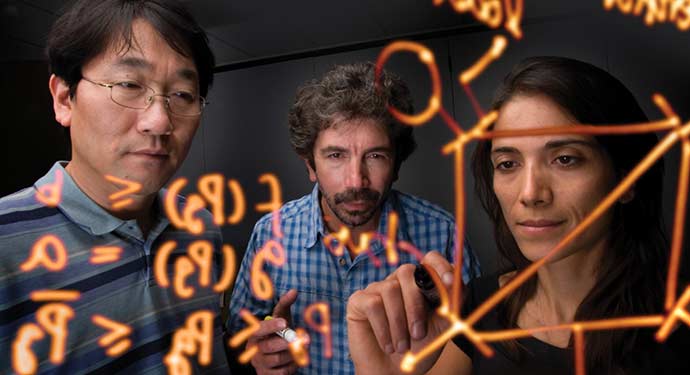 A photo of a group of people writing equations in orange marker on a clear, plastic board.