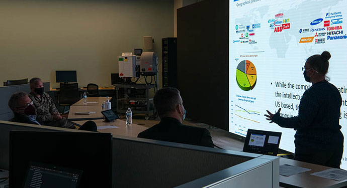 A woman presents in front of a screen to three people in medical masks.