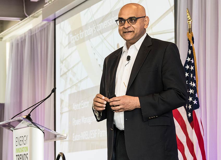 Photo of a man wearing a business suit and standing on a stage with an American flag, projector screen, and podium behind him.
