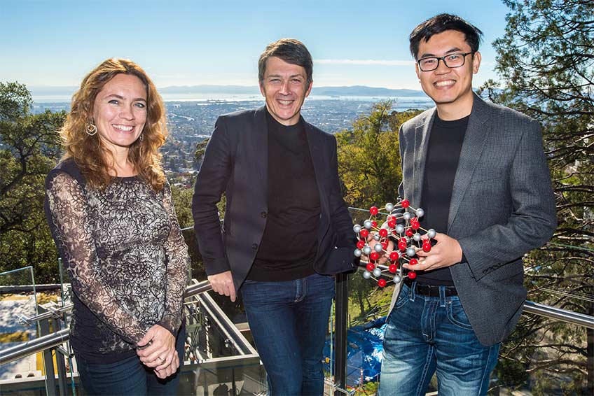 One woman and two men stand outside on a balcony. The man on the right holds a ball-and-stick model of a molecule.