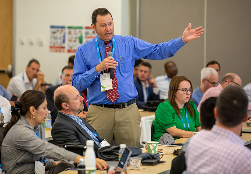 Photo of a man standing in a seated crowd while holding a microphone to speak