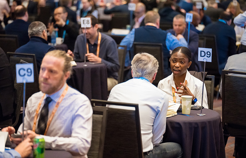People in a hotel ballroom meet at dozens of small tables talking with each other.