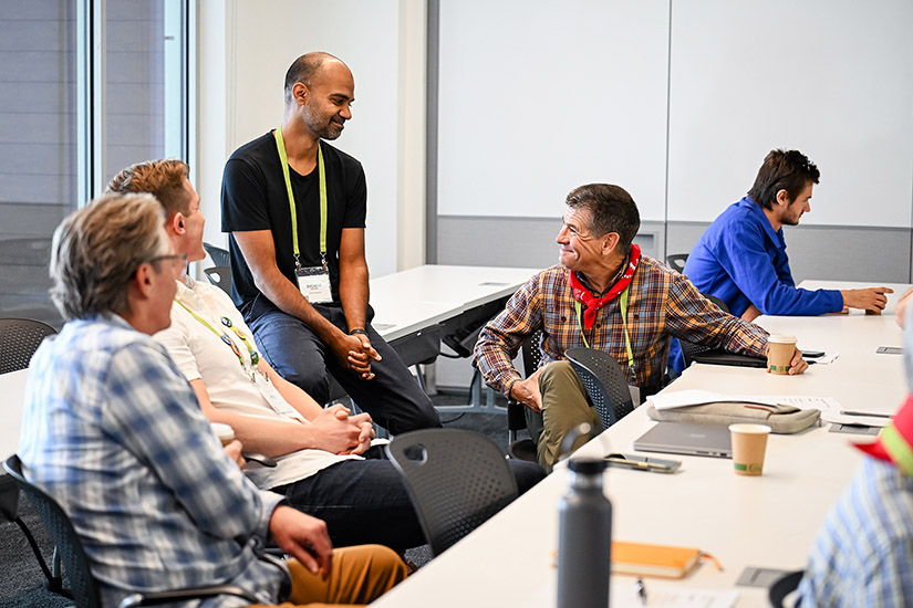A man in a black shirt speaks with other men who are sitting in a classroom.