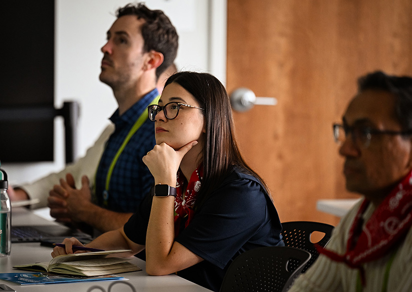 A woman in glasses sits between two men in a classroom watching a speaker. 