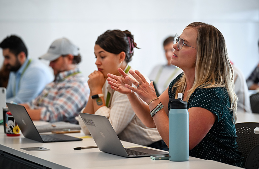 A woman sitting at a long table talks to the person in the front of a classroom, with another woman and two men in the background.