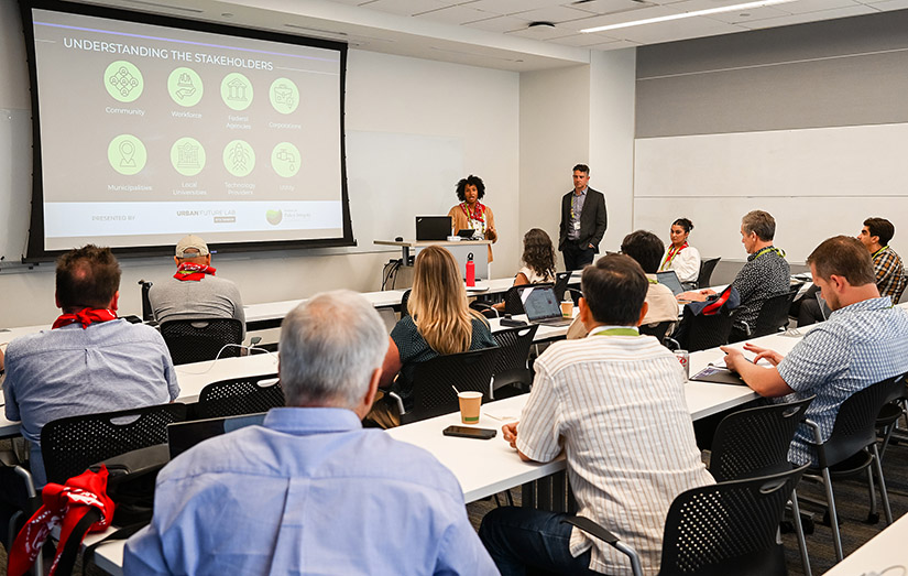  Two people speak before a full classroom next to a screen.