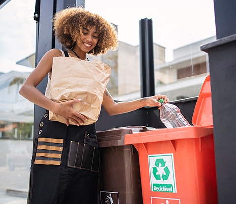 A woman holding a paper bag places plastic bottles into a recycling bin.