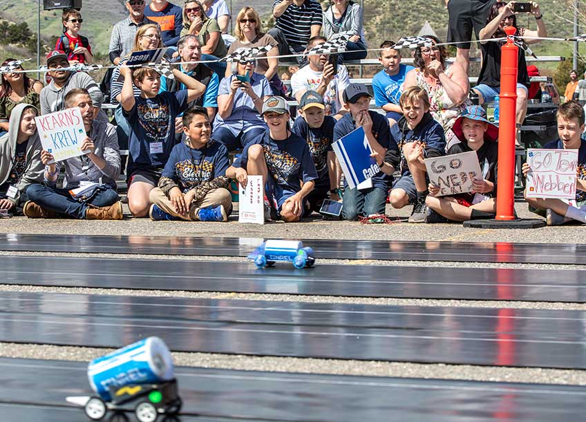 A group of students and parents hold signs as they cheer on model cars racing down a track.