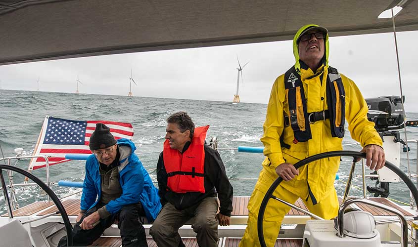Three people on a boat driving away from wind turbines in the ocean.