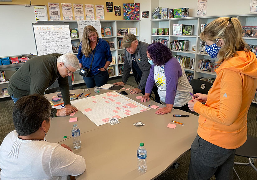 Several people stand around a table examining a large white sheet of paper with pink labels on it.