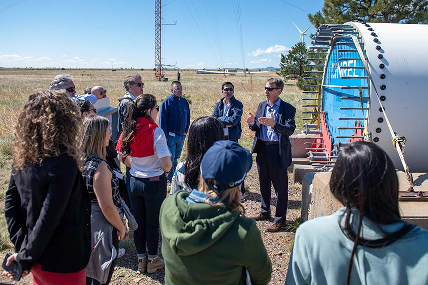 A man presents to a group of people outdoors while standing in front of wind turbine equipment.