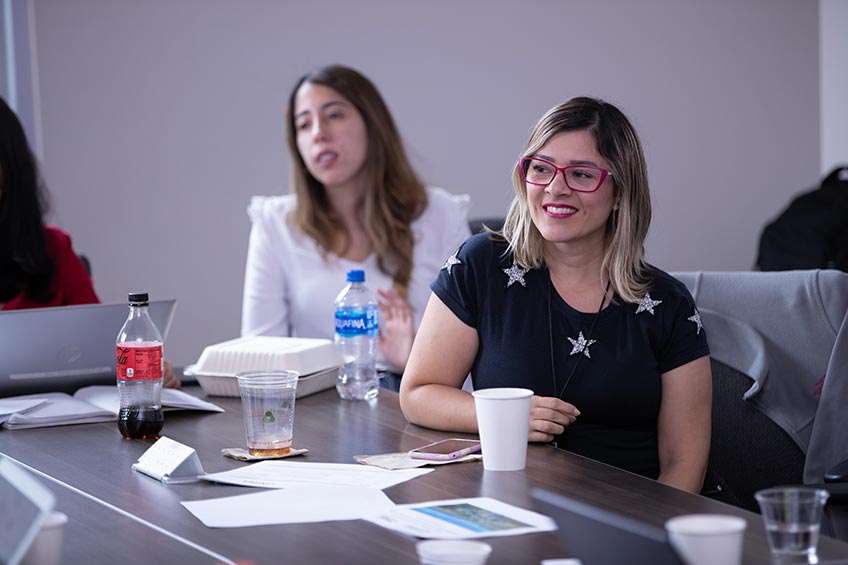A seated women is smiling. There is another women present in the background.
