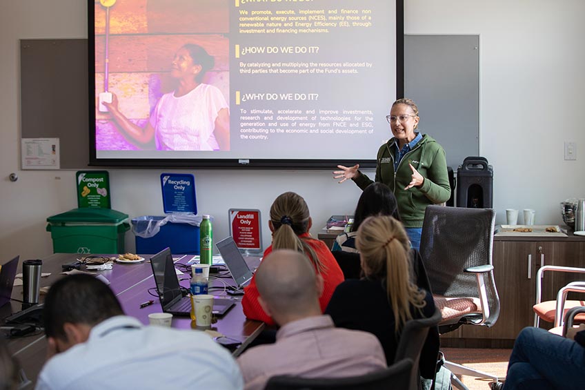 A women stands at the front of a conference room with a presentation behind him. He is gesturing at a group of seated people.