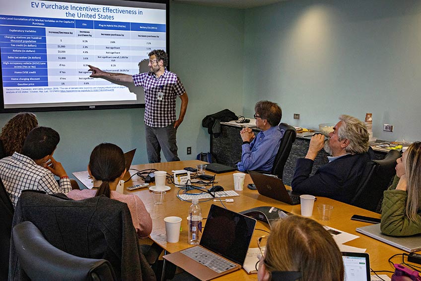 A man stands at the front of a conference room with a presentation behind him. He is gesturing at a group of seated people.