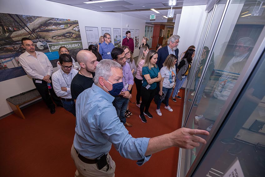 A man is pointing to something out of site as a group of people with him look through glass at a facility on the other side.