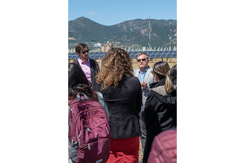 A man is presenting in front of a group of people outdoors. There are solar PV panels in the background.