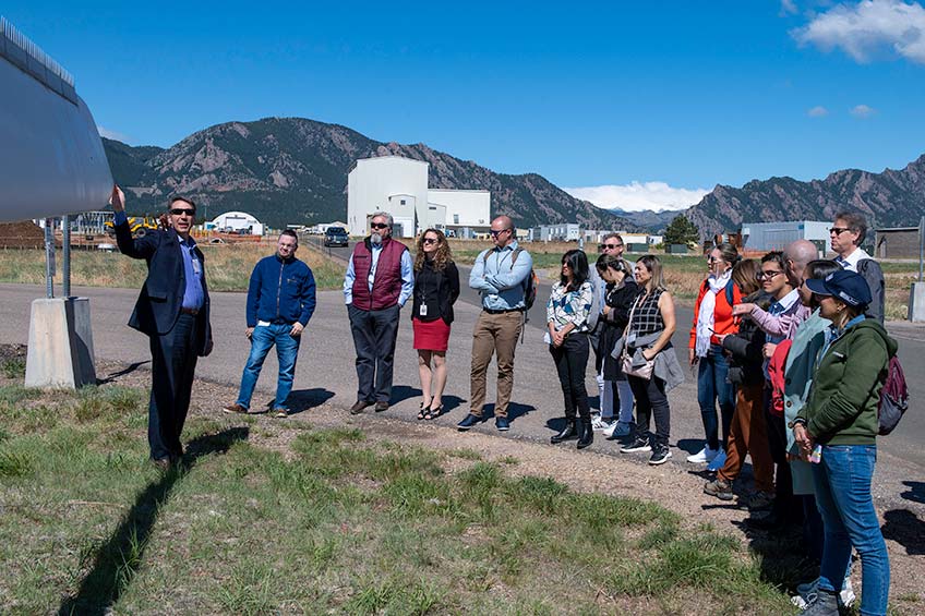 A man is presenting in front of a group of people outdoors. There is a wind turbine in the background.