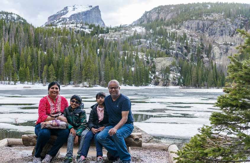 Mou Paul, her husband, and twin sons sit on a log bench in front of an icy lake surrounded by mountains.