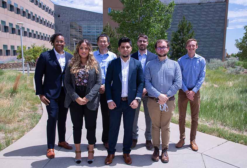 Seven people in business attire standing on a sunny sidewalk outside a building