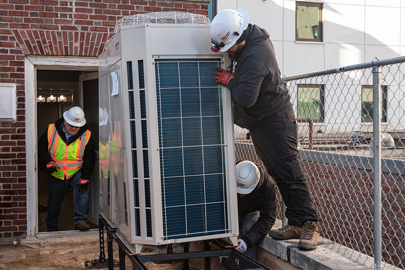Three men work on installing an HVAC device that looks like a phone booth with a fan on top.
