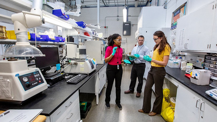 Three people standing in a laboratory wearing safety goggles and latex gloves