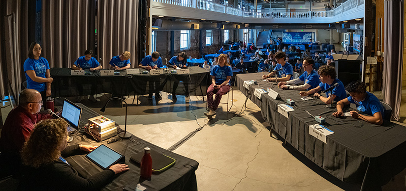 Two tables of students face a table of judges on a stage.