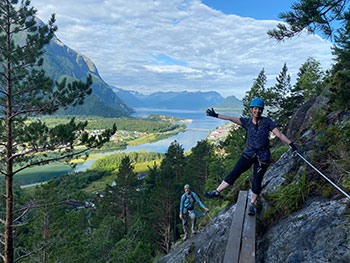 A person in a helmet stands on a wooden plank attached to the side of a rock. One person stands below her with a river and mountains behind.