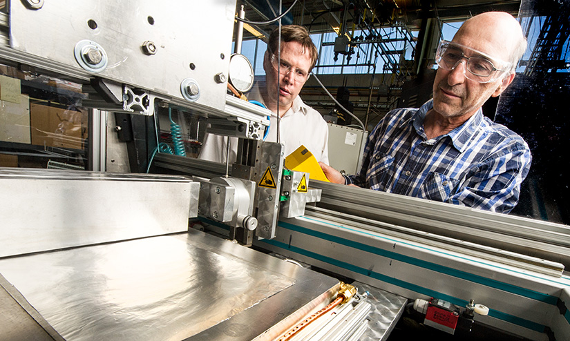 Two men wearing safety goggles use a machine that has a paper-thin piece of metal on it.