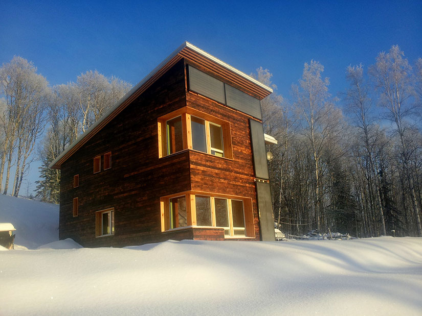 A modern home with dark wood siding and large windows with a snow-covered lawn