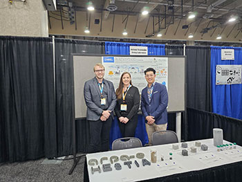 Aday and two other people standing in a conference booth with a table holding small pieces of cement