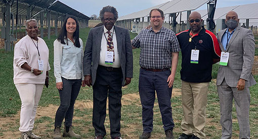 Six people stand in front of commercial-scale solar panels on a farm.